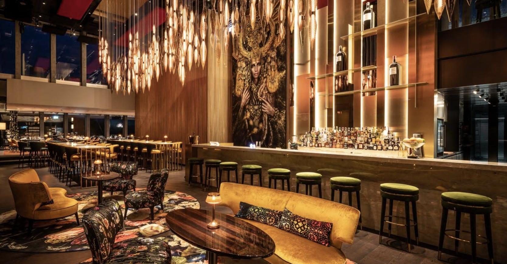 Die Lounge-Bar des Capsule Hotels hat goldene Kronleuchter, ein Wandbild, beleuchtete Regale, grüne Barhocker und gemütliche florale Sitzgelegenheiten.
