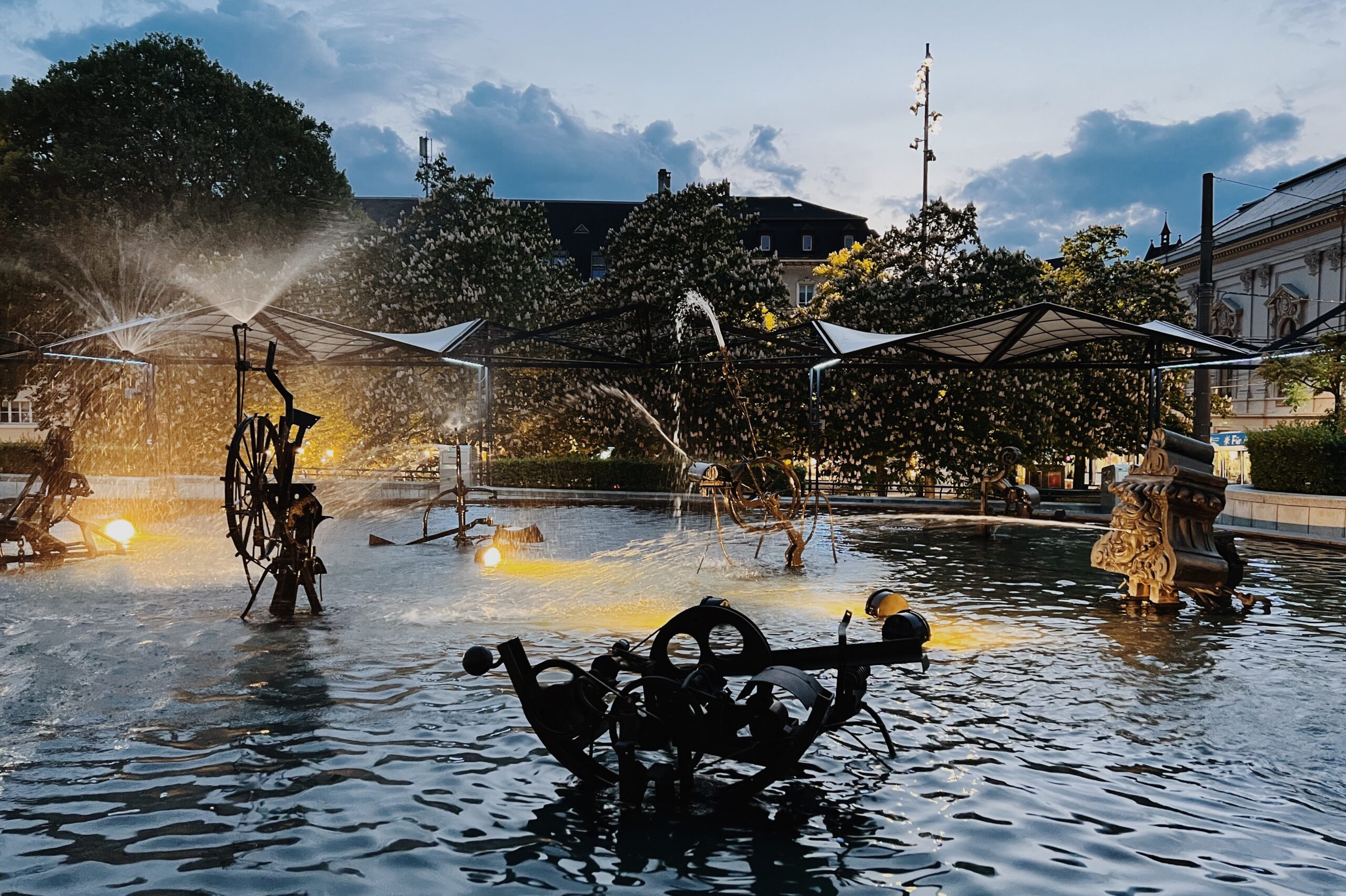 Der Tinguely-Brunnen in Basel in der Abenddämmerung, mit kinetischen Metallskulpturen, die Wasser über ein beleuchtetes Becken spritzen