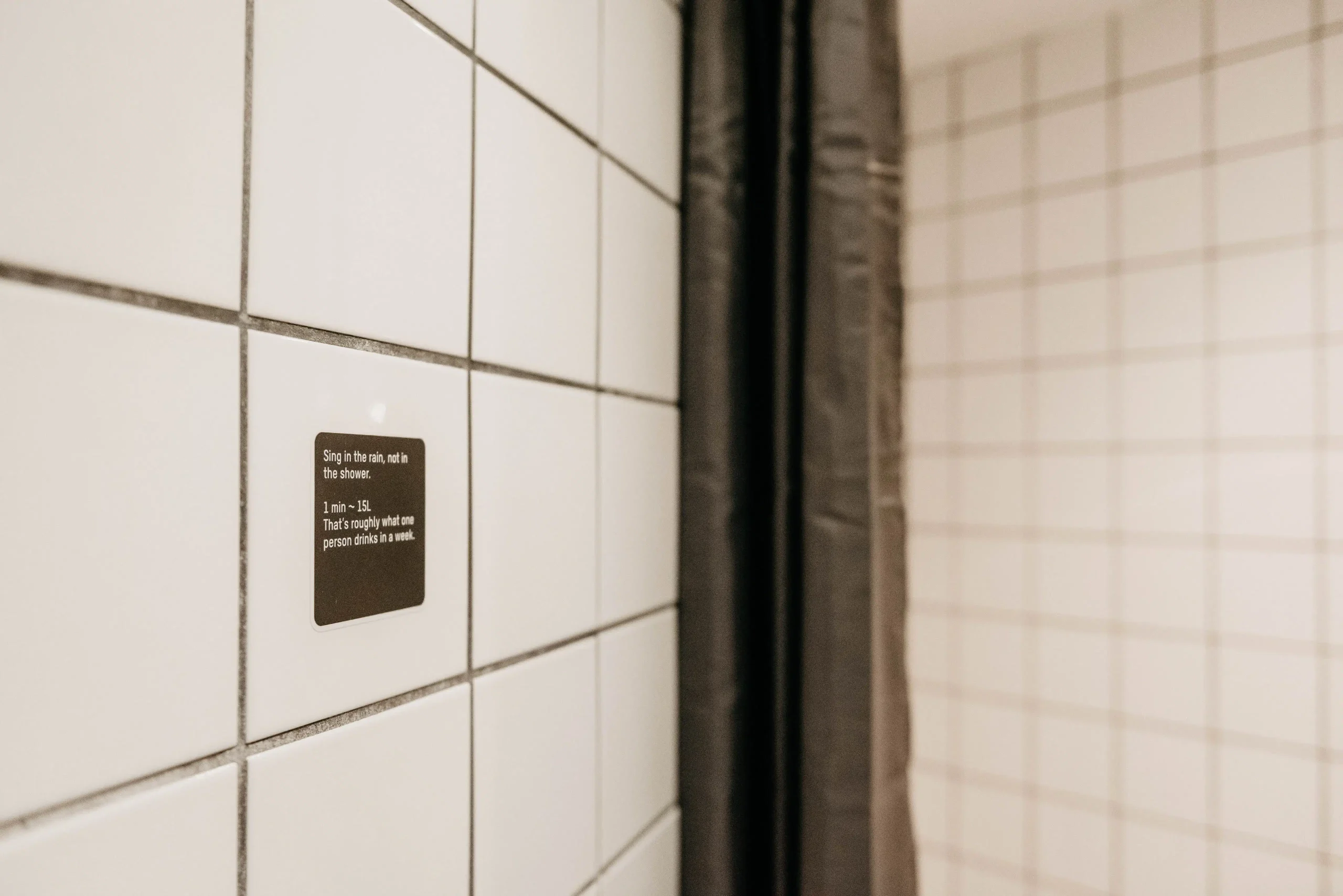 White tiled wall with an eco-friendly shower reminder at Capsule Hotel Copenhagen Vesterbro.