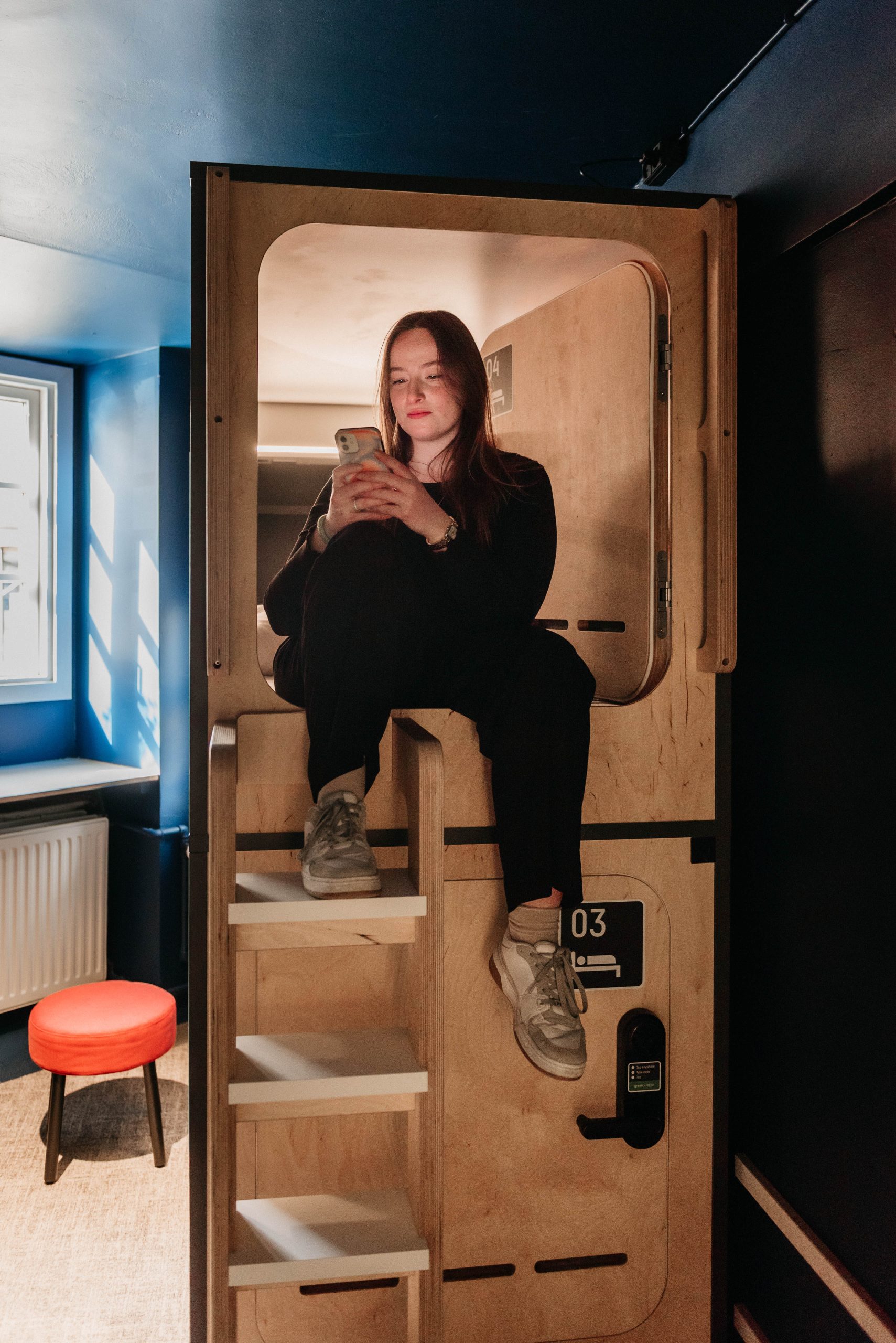 Person sitting at capsule entrance, Capsule Hotel Copenhagen Vesterbro, with cozy interior and ladder access.