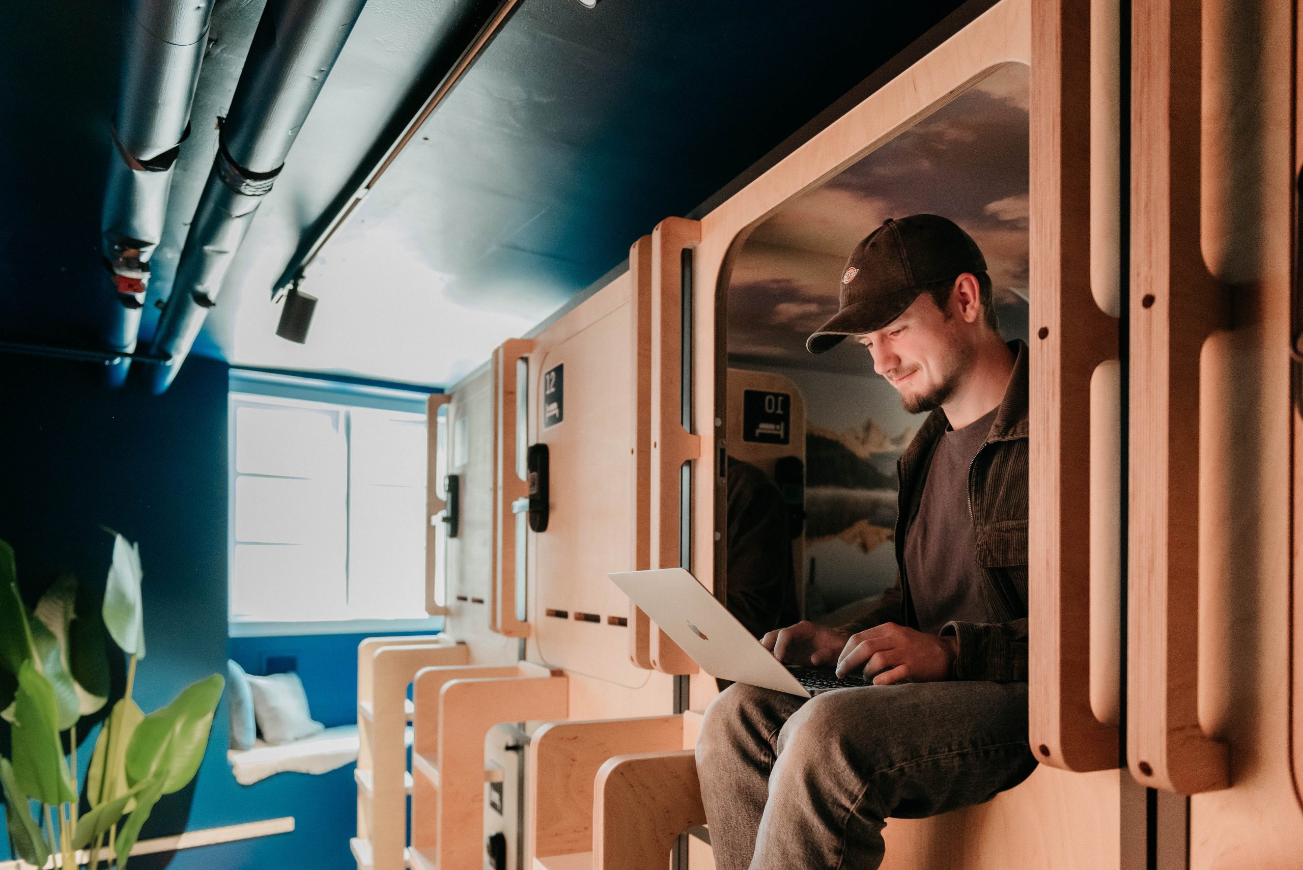 Man working on laptop at Capsule Hotel Copenhagen Vesterbro, seated on capsule bed in modern dorm setting.