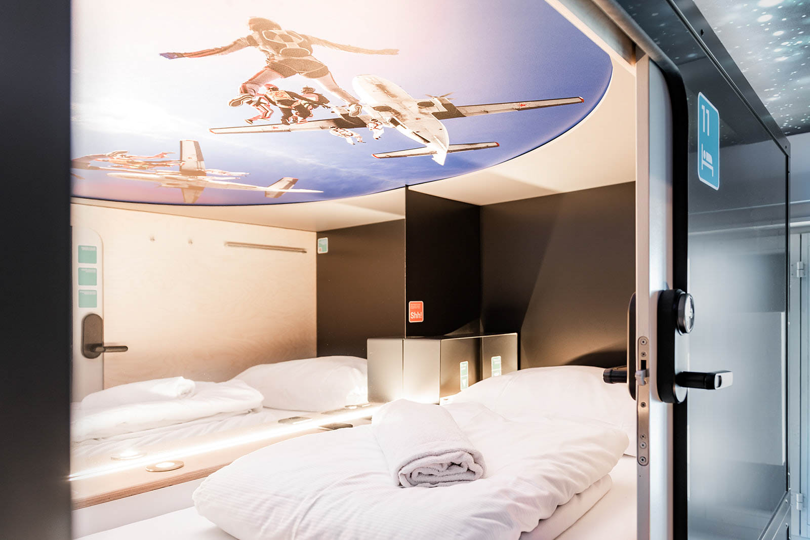 Capsule Hotel sleeping pod with white bedding and rolled towel, skydiver ceiling mural, soft LED lighting
