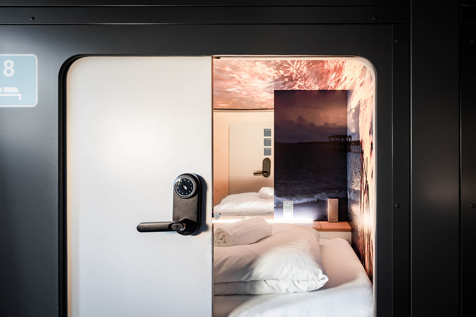 Comfortable capsule room with beach artwork at Capsule Hotel Lucerne The Lab. Cozy bedding and illuminated entry.