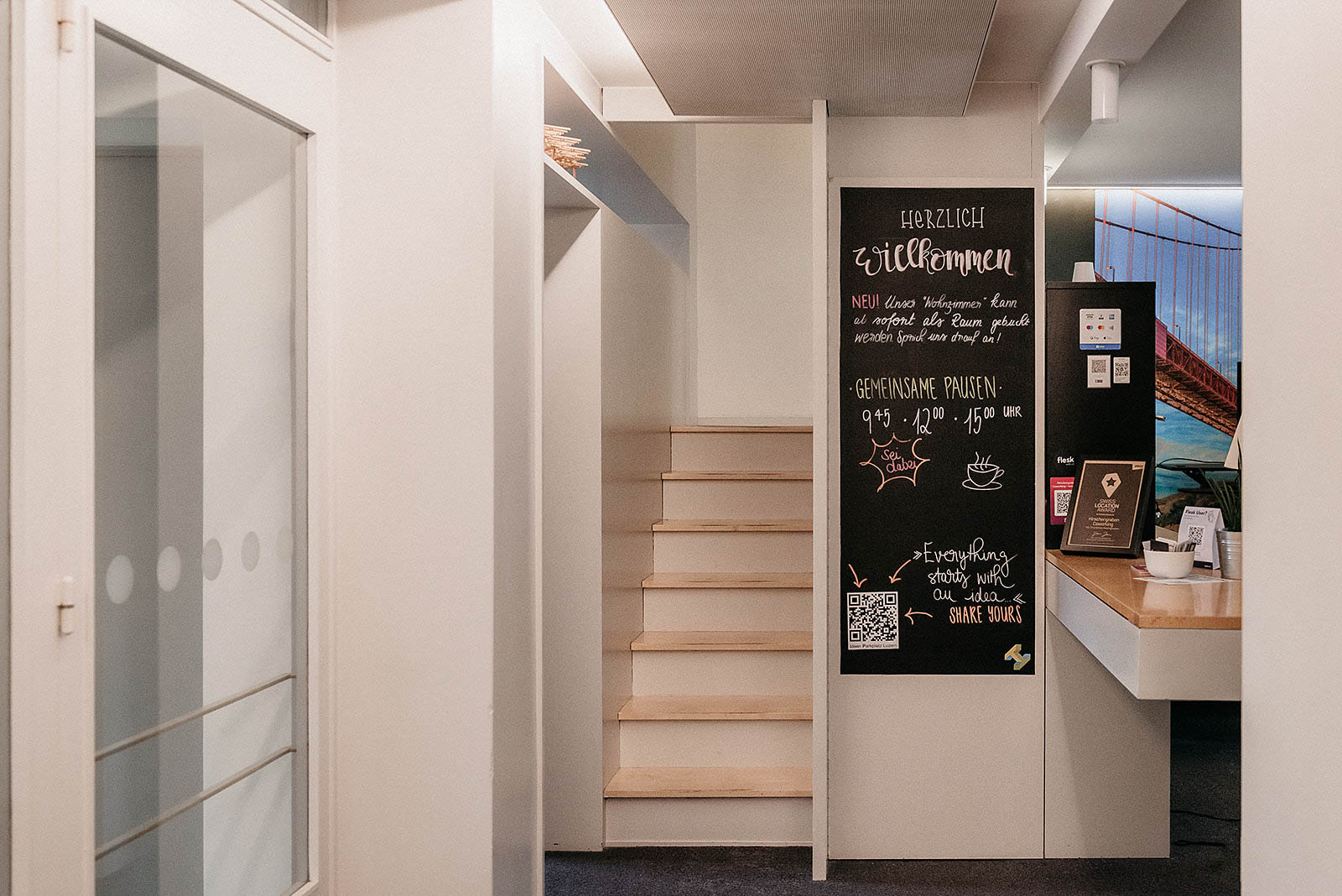 Capsule Hotel minimalist hallway with light wood stairs, welcome chalkboard, and reception desk by Golden Gate bridge mural.