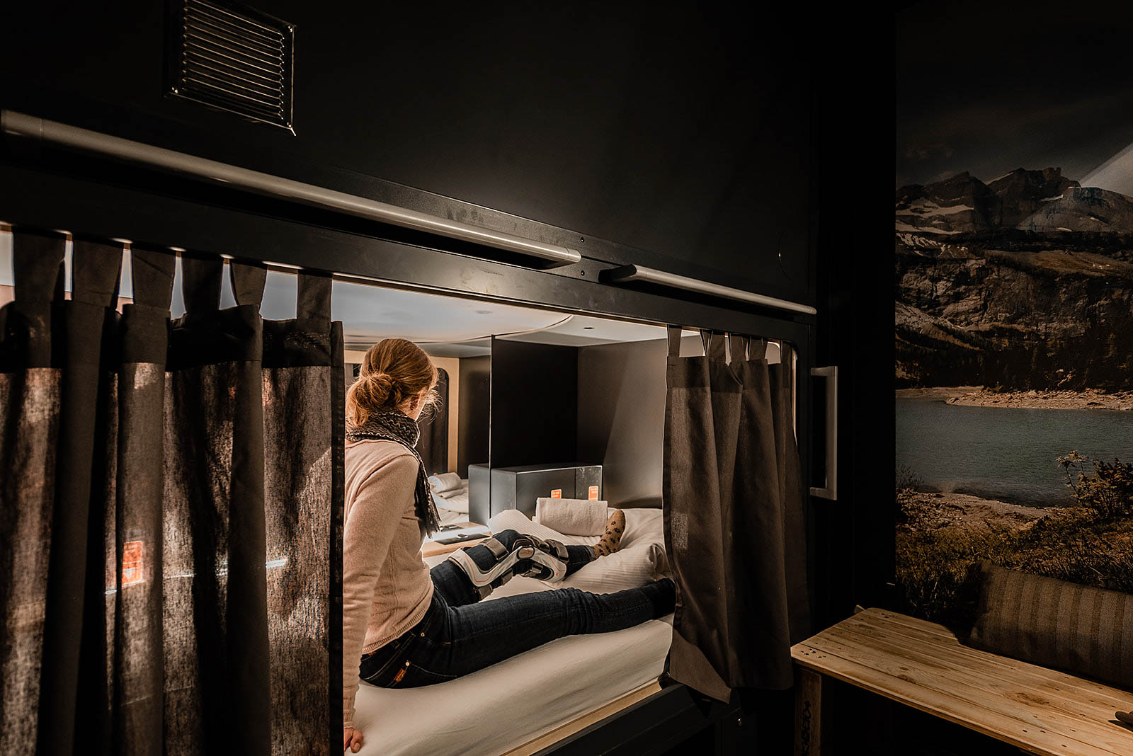 Woman relaxing in a capsule room at Capsule Hotel Alpine Garden Zurich Airport with mountain mural and ambient lighting.