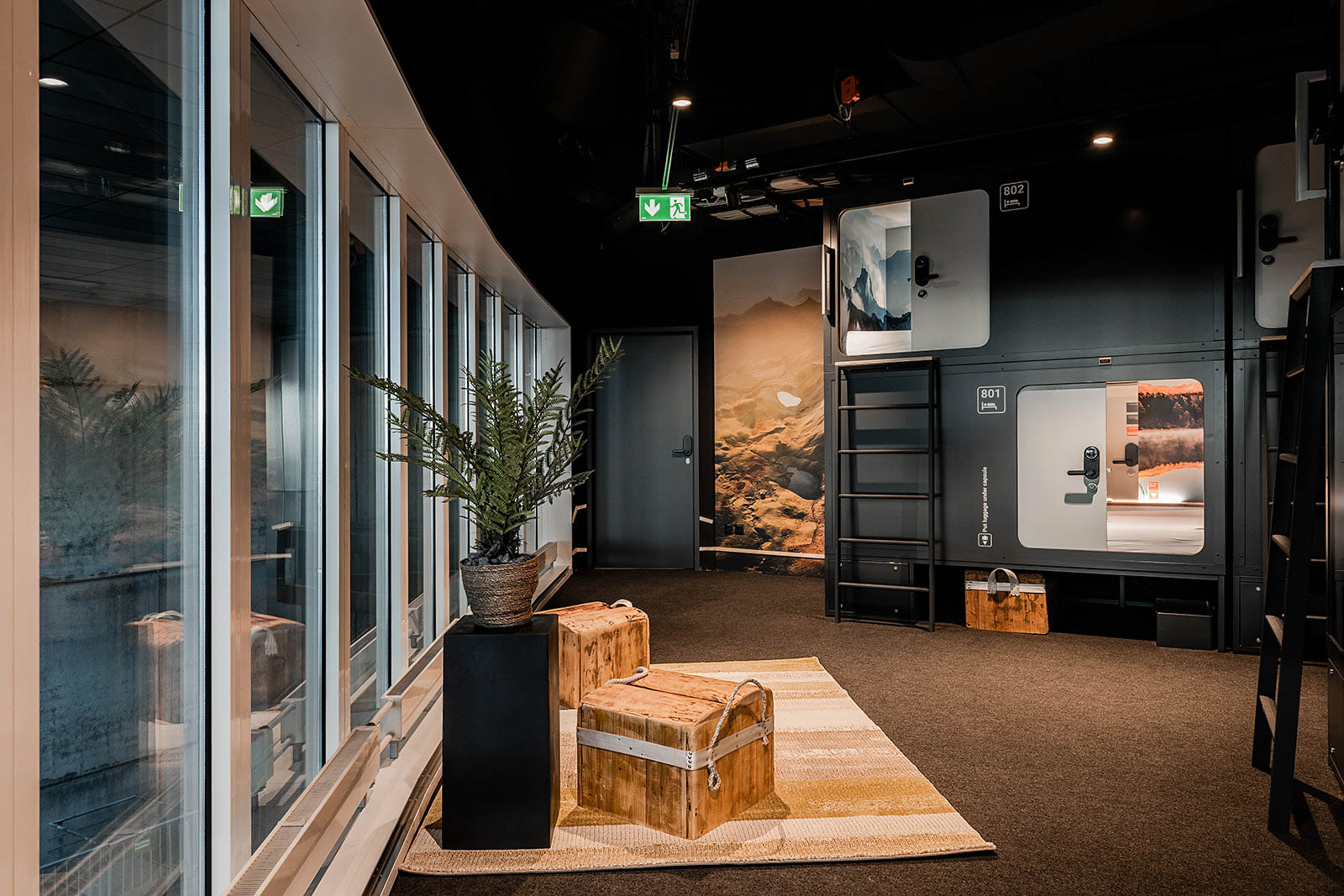 Spacious hallway with capsule rooms, seating, potted plant, and scenic wall art at Capsule Hotel Alpine Garden Zurich Airport.