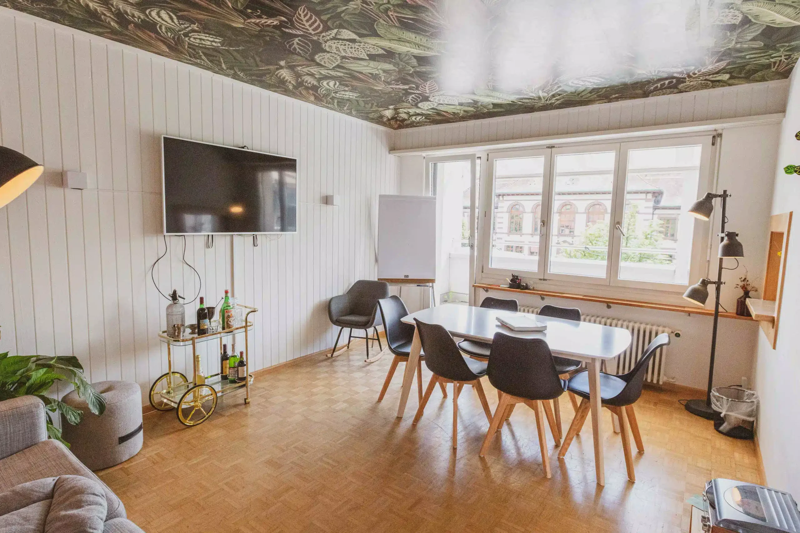 Meeting-Lounge im Capsule Hotel mit Tisch, schwarzen Stühlen, Wandfernseher, Barwagen und grossen Fenstern unter einer begrünten Decke.