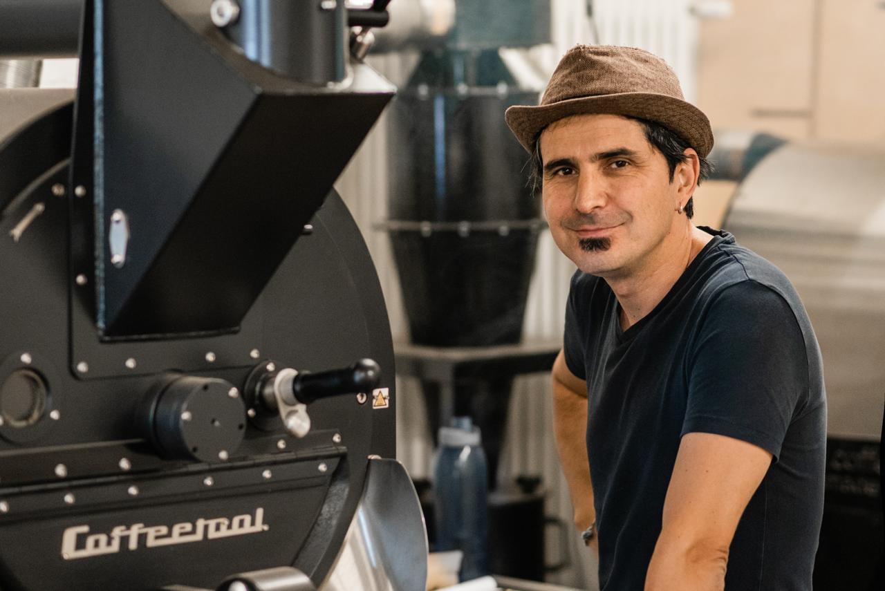 Capsule Hotel team member in a brown hat, smiling beside a large coffee roaster in the hotel café.