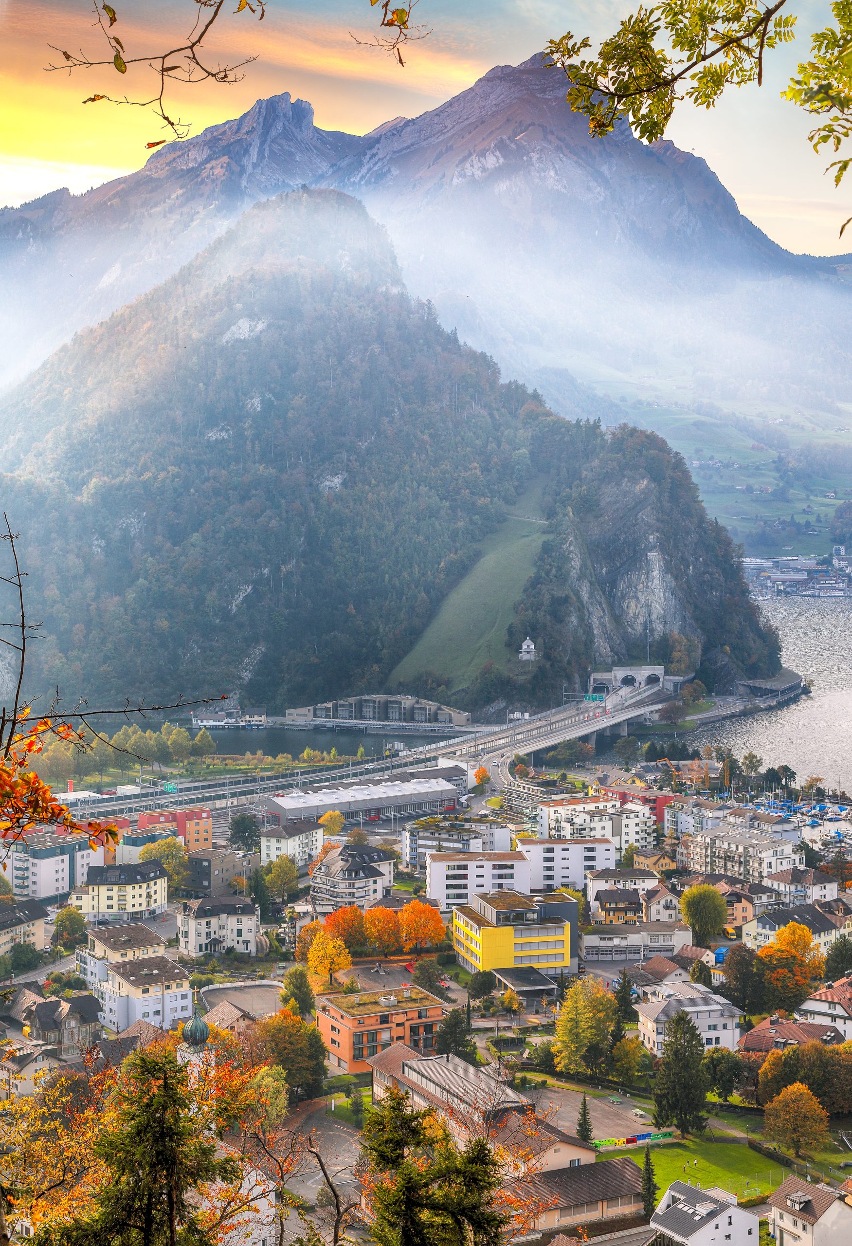 Nebelverhangene europäische Alpen über einer farbenfrohen Stadt am See bei Sonnenaufgang, Herbstbäume und Autobahntunnel, Capsule Hotel.
