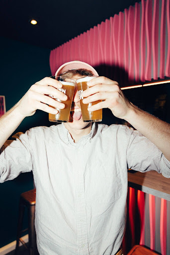 Ein Mann mit zwei Bieren bedeckt spielerisch seine Augen in einer lebhaften Capsule Hotel Lounge mit farbenfroher Einrichtung.