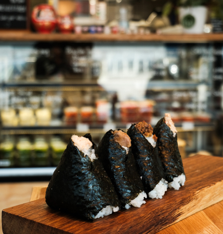 Onigiri-Reisdreiecke auf einem Holzbrett im Café des Capsule Hotels, im Hintergrund verschwommen ein Deli-Kühlschrank.