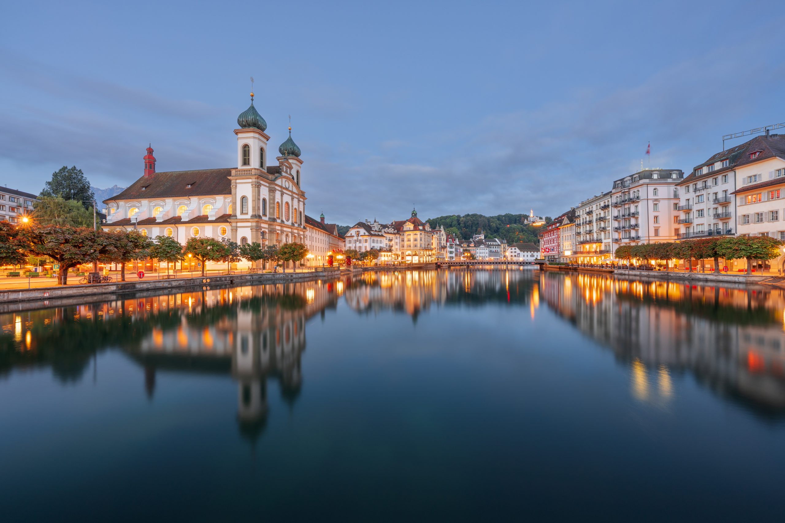 Ein Markenbild vom Capsule Hotel: eine europäische Flusslandschaft in der Dämmerung mit einer Kuppelkirche und Lichtern, die sich auf dem ruhigen Wasser spiegeln.
