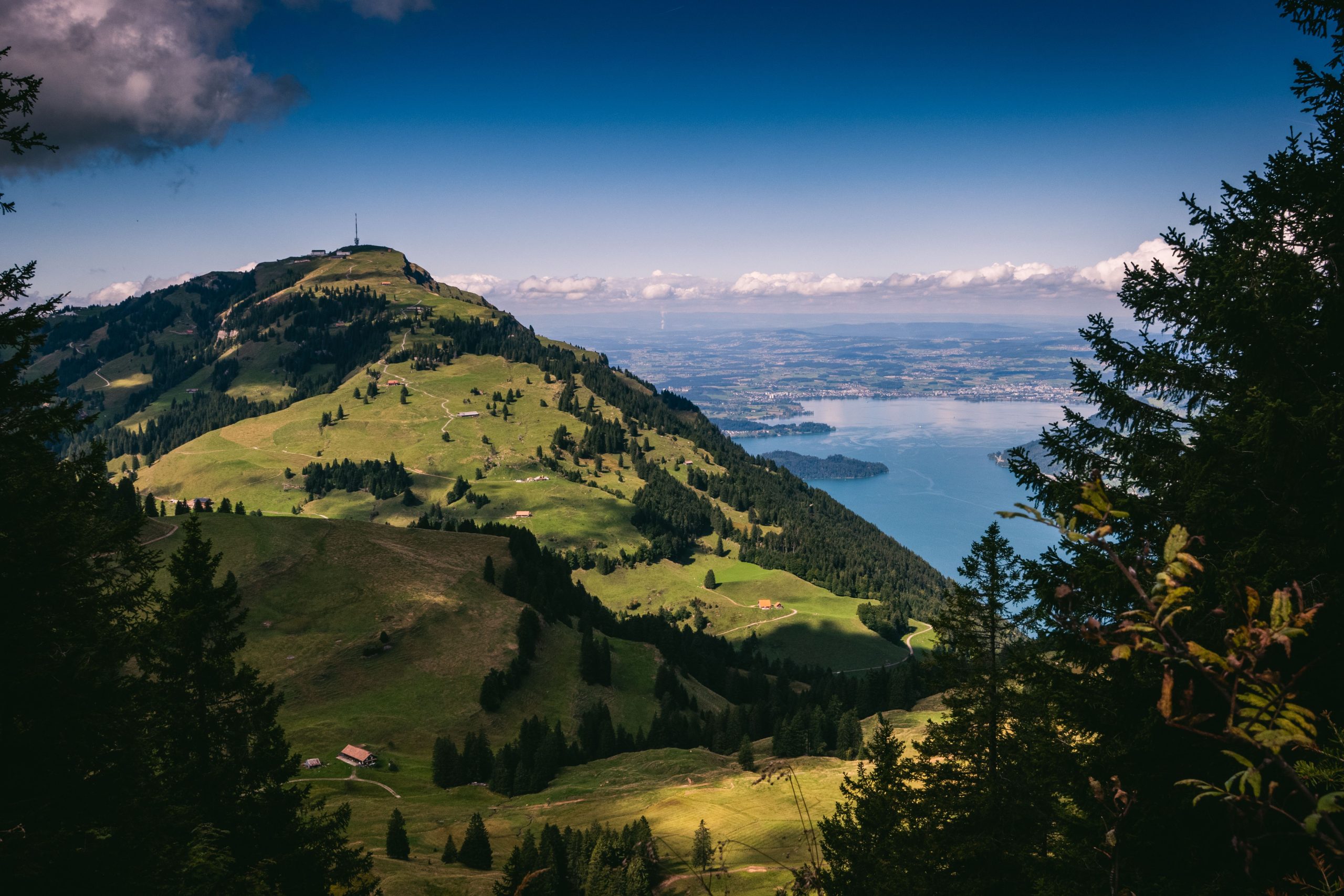 Capsule Hotel Alpenlandschaft: grüner Bergrücken über einem blauen See, Wanderwege und Chalets unter einem hellen Himmel, umrahmt von Tannen.