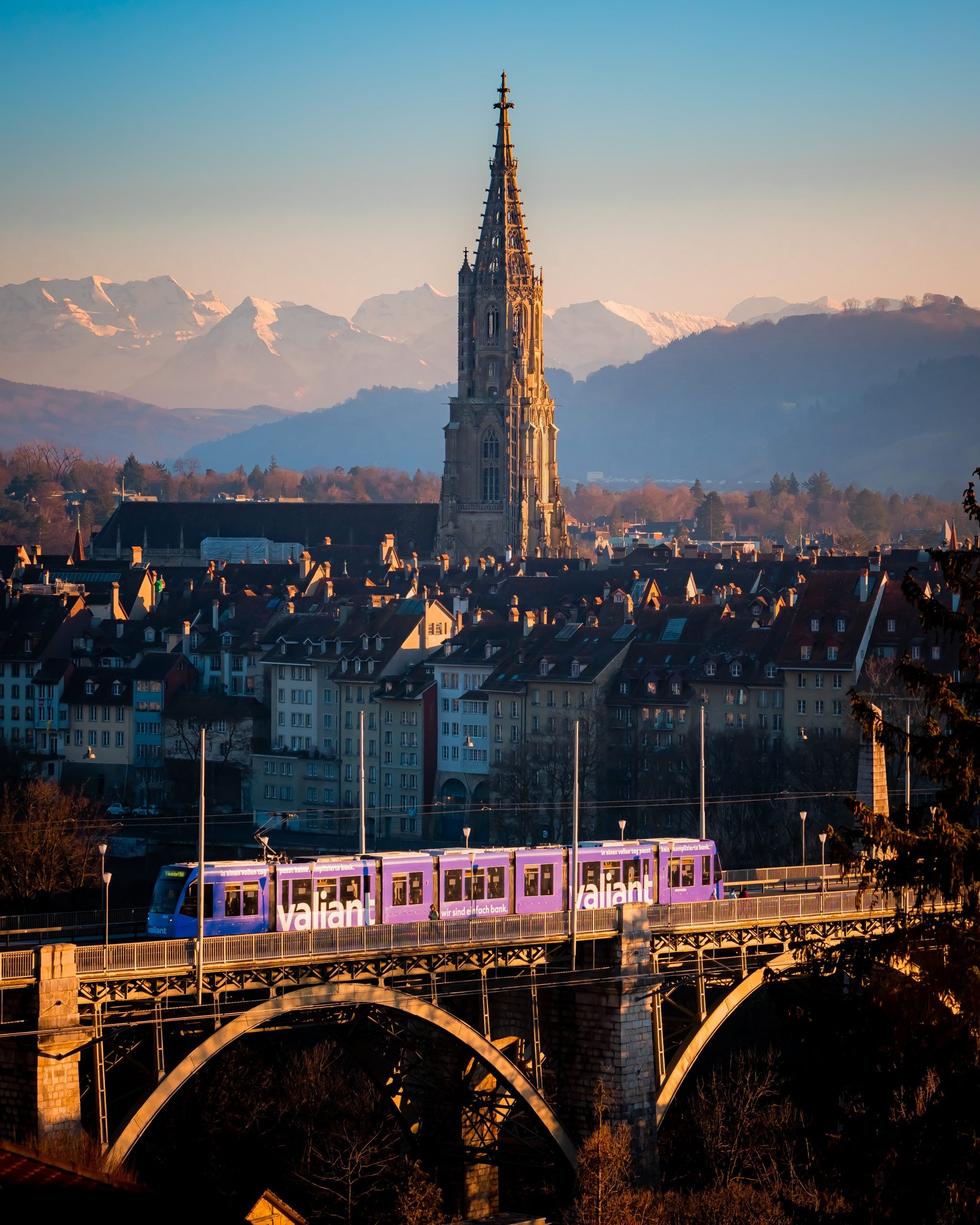 Capsule Hotel tram crossing a stone bridge; historic rooftops, cathedral spire, and snow-capped mountains at dusk.