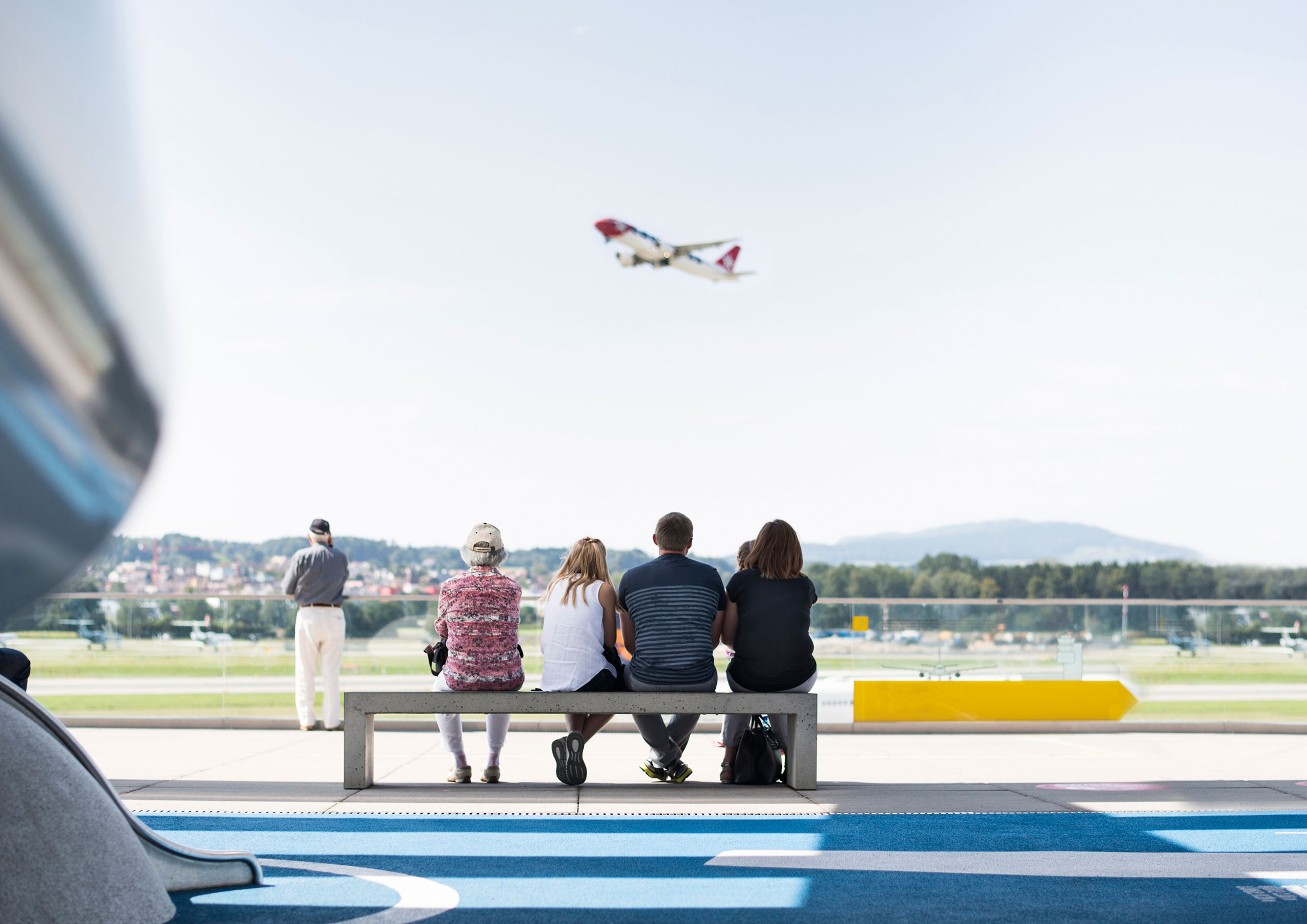 Reisende des Capsule Hotels beobachten von einer Flughafenterrasse aus, wie ein Flugzeug startet, sitzend auf einer Bank mit Blick auf die Startbahn.