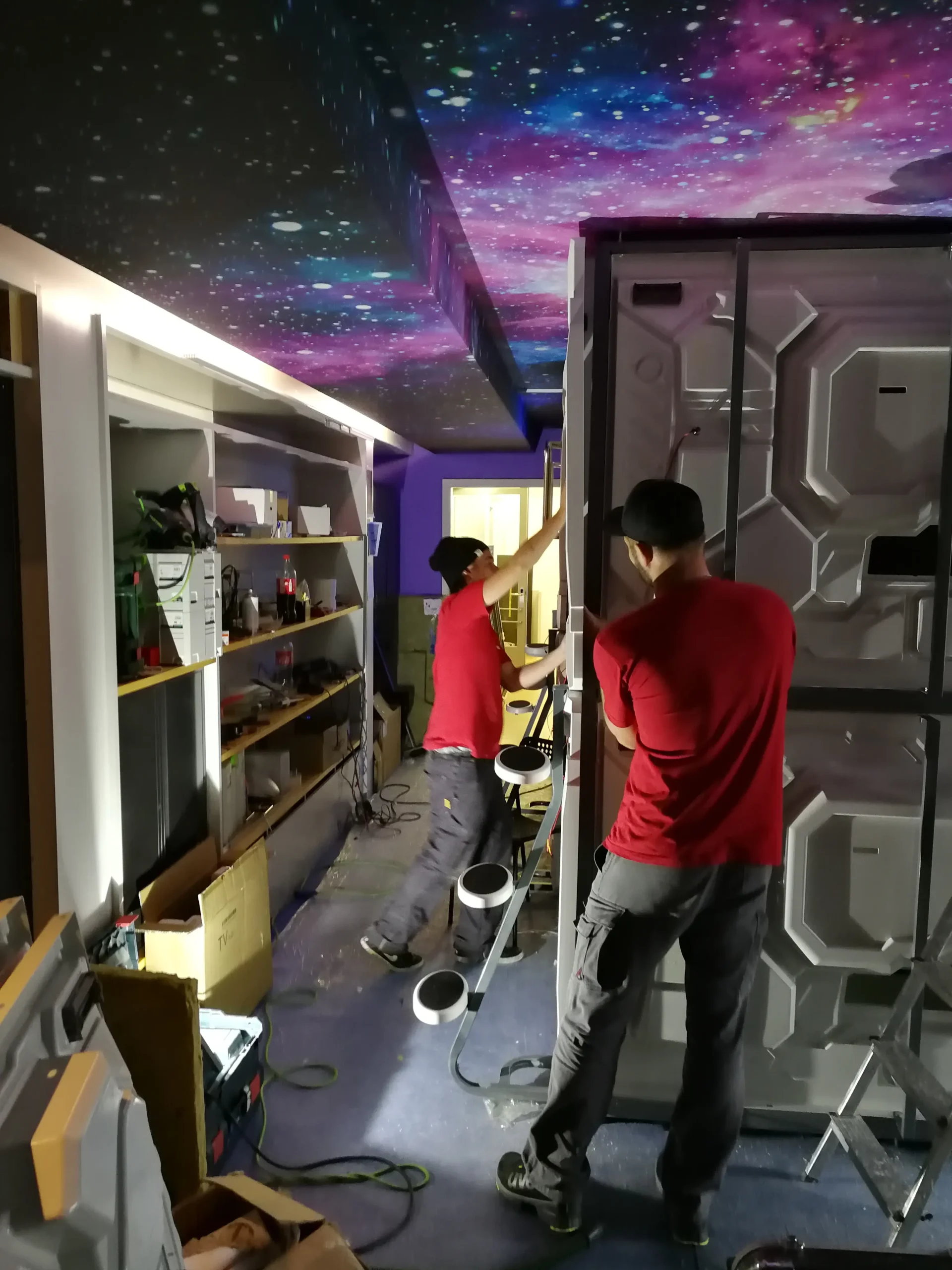 Two workers in red shirts installing a capsule pod under a galaxy-themed ceiling at Capsule Hotel.