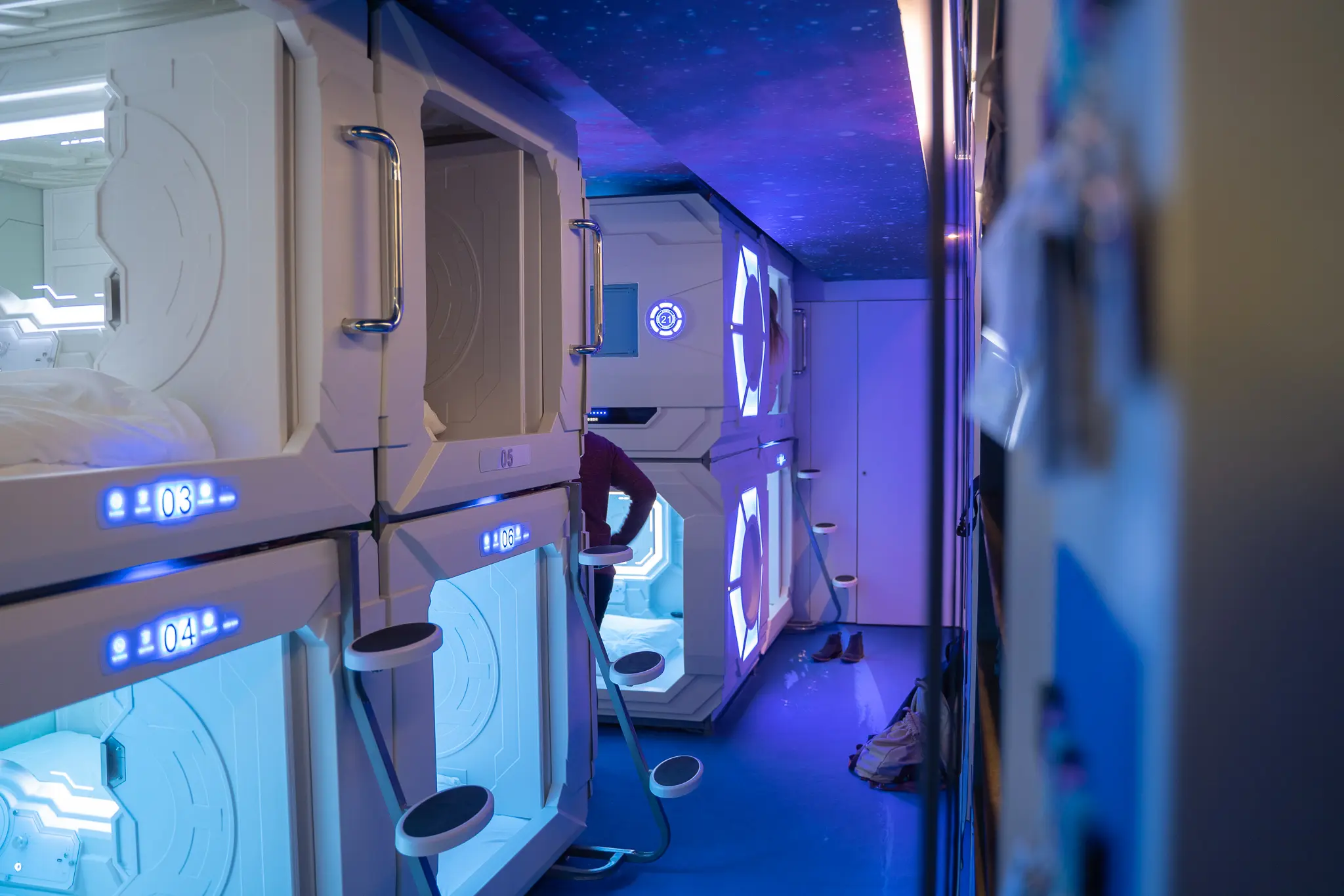 Spacious sleeping pods with blue lighting at Capsule Hotel, featuring futuristic design and cozy amenities.