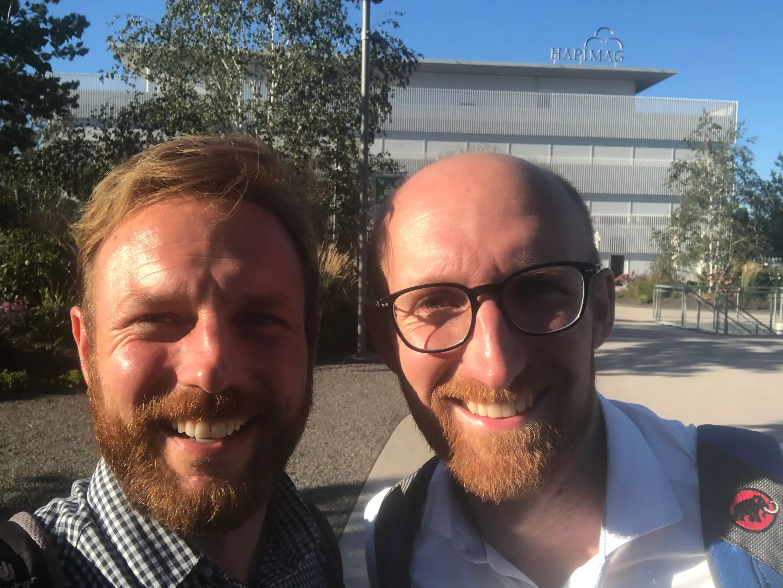 Two smiling team members outdoors, showcasing camaraderie at Capsule Hotel. Building with trees in the background.