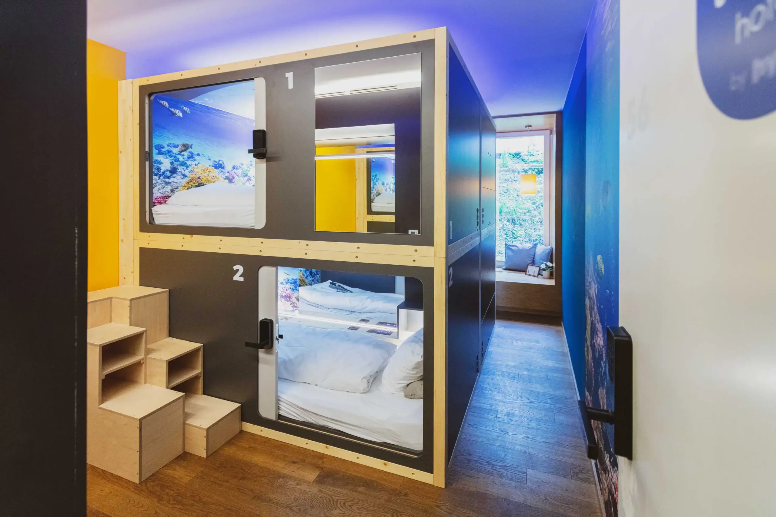 Two stacked sleeping pods in a vibrant hallway at Capsule Hotel, showcasing modern, cozy design and colorful decor.