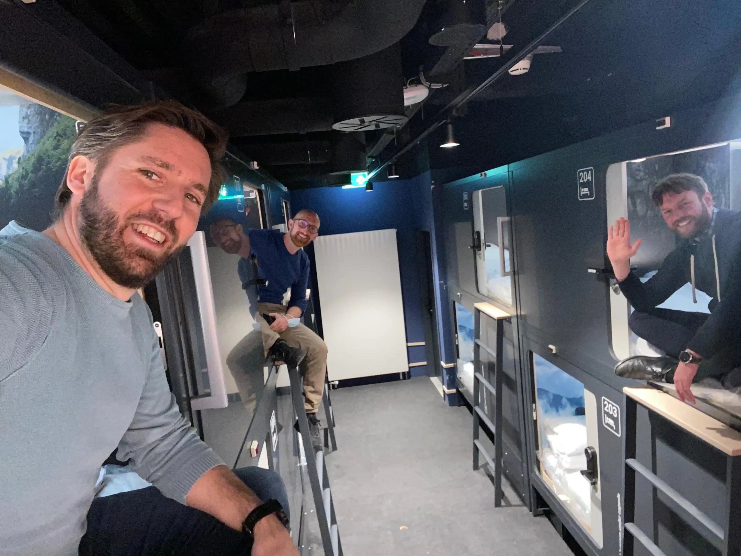 Three team members smiling and sitting near capsule units at Capsule Hotel, showcasing modern, cozy design features.