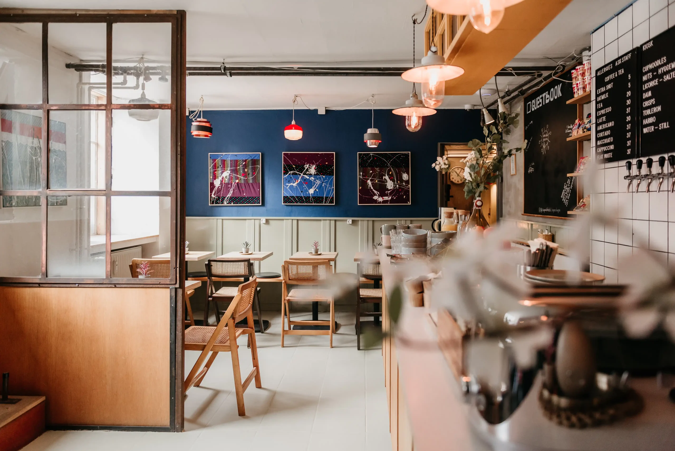 Capsule Hotel café with wooden chairs, pendant lights, blue wall art, and tiled bar with taps.
