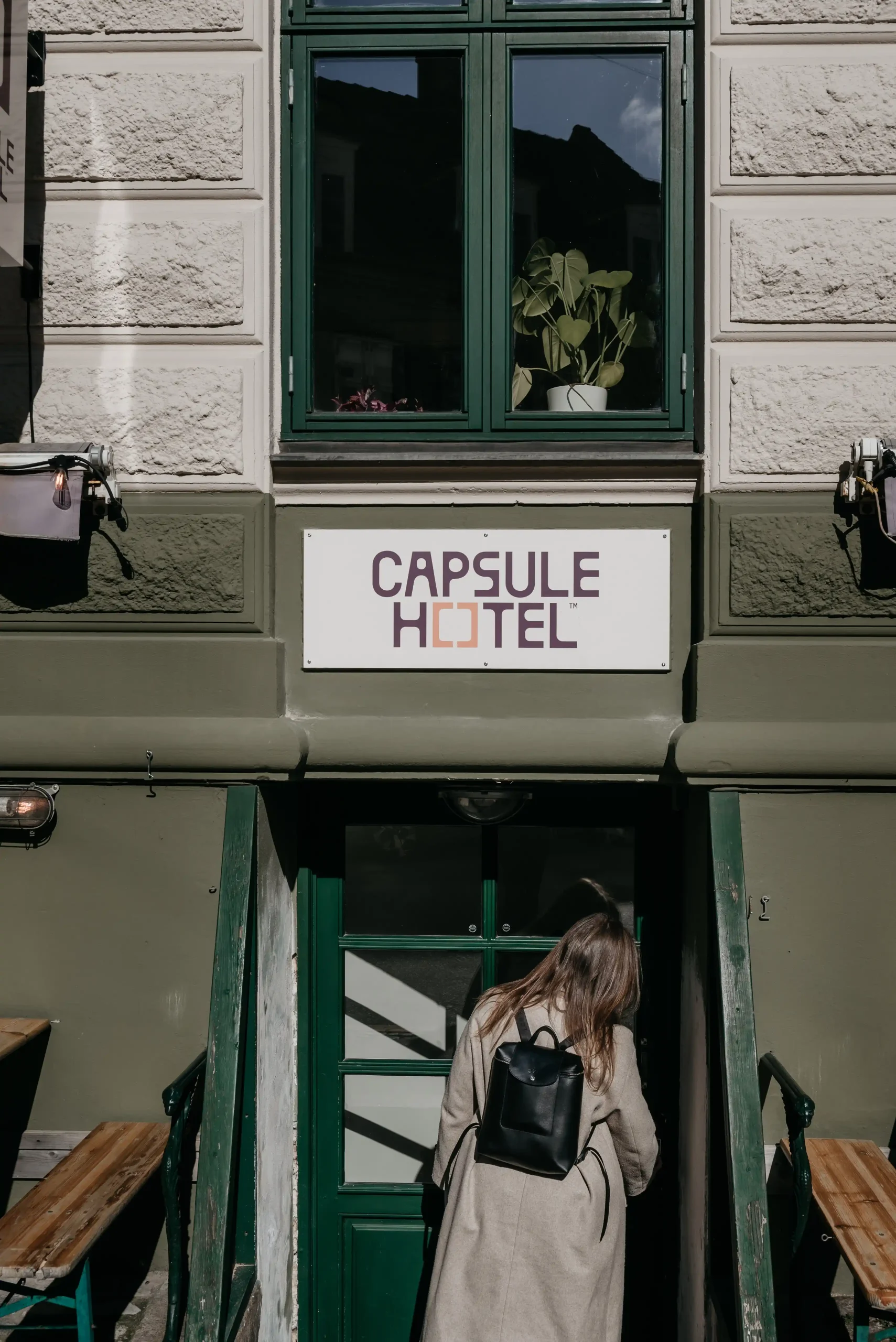Guest with black backpack entering green door at Capsule Hotel entrance on historic European facade.
