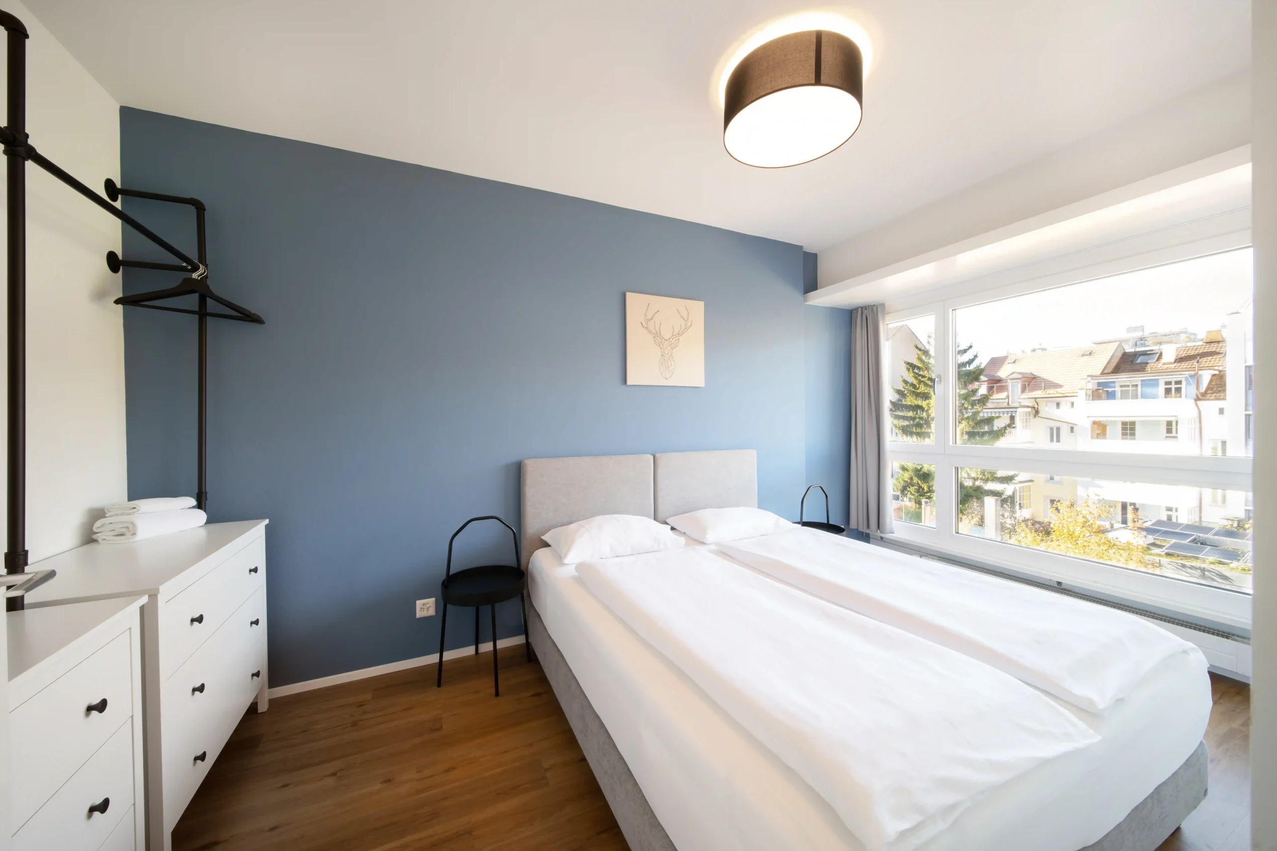 Stylish private apartment at Hyve Basel Capsule Hotel featuring a double bed, blue accent wall, and large window view.