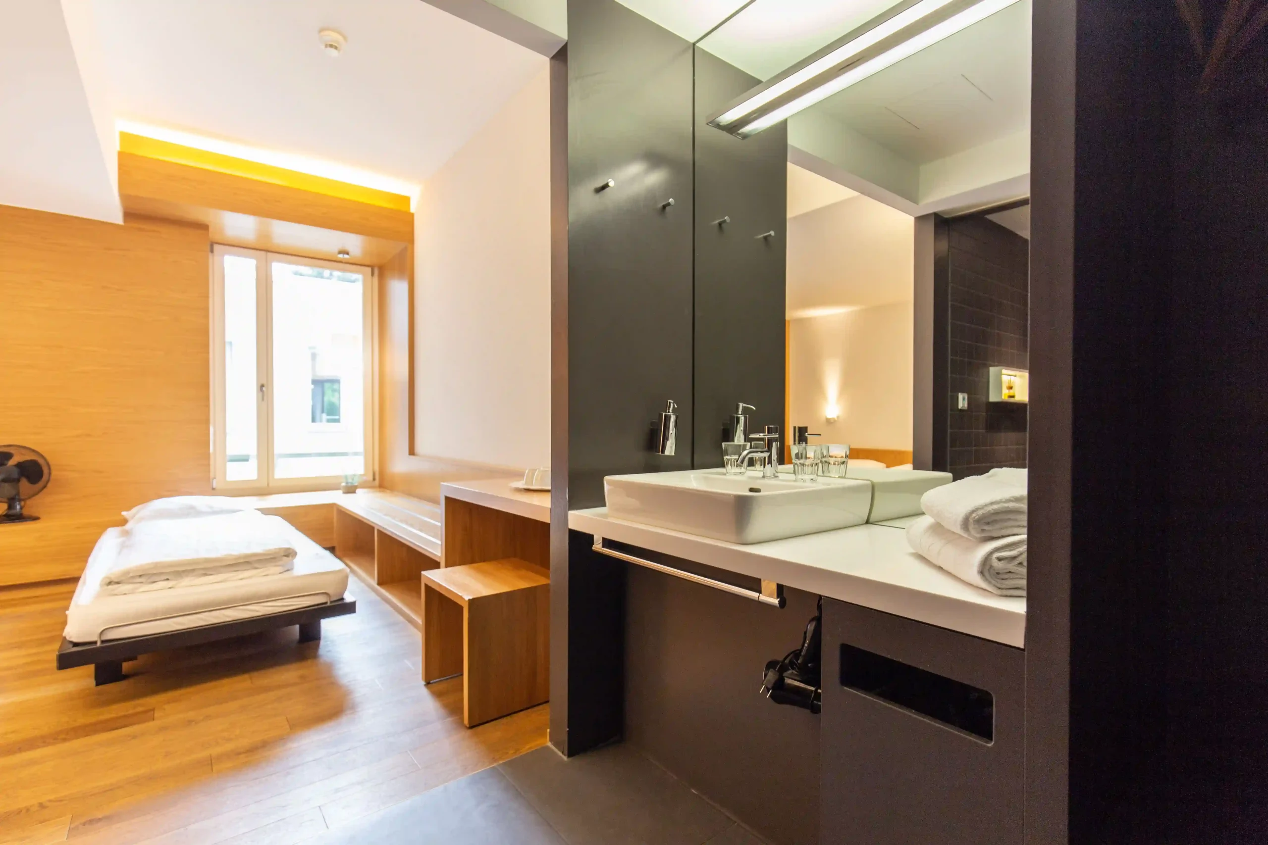 Sleek Capsule Hotel room featuring modern bed, wooden accents, and a minimalist bathroom with large mirror.