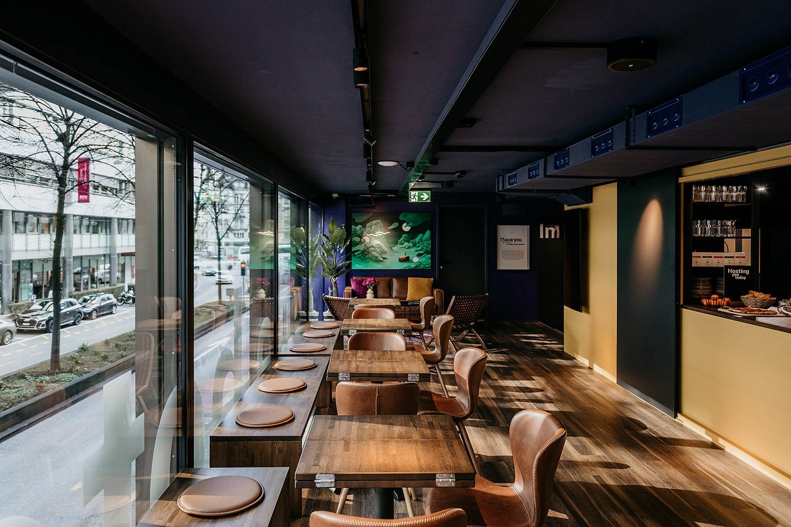 Dining area with tables and chairs at Capsule Hotel Lucerne Old Town, overlooking city street through large windows.