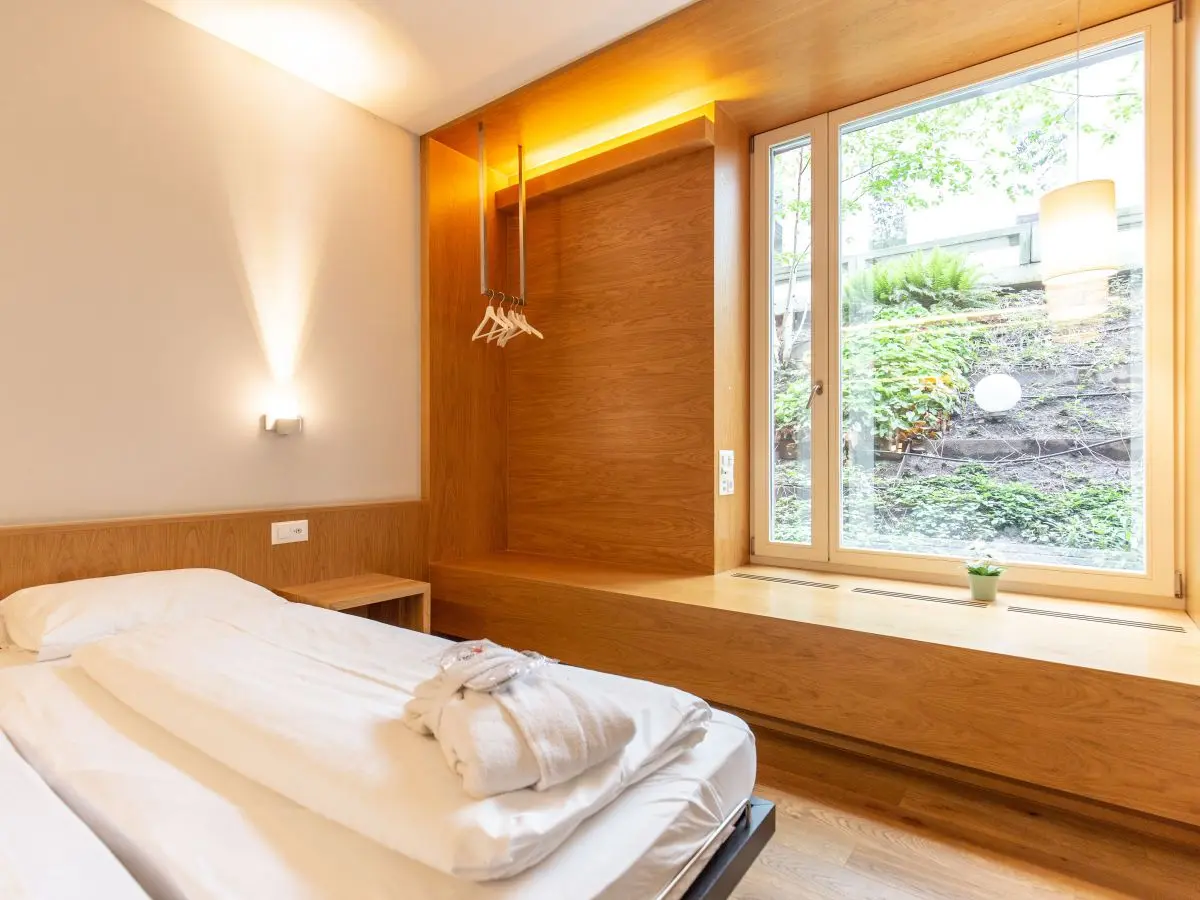 Cozy Capsule Hotel room featuring a neatly made bed, wooden decor, and a large window with a garden view.