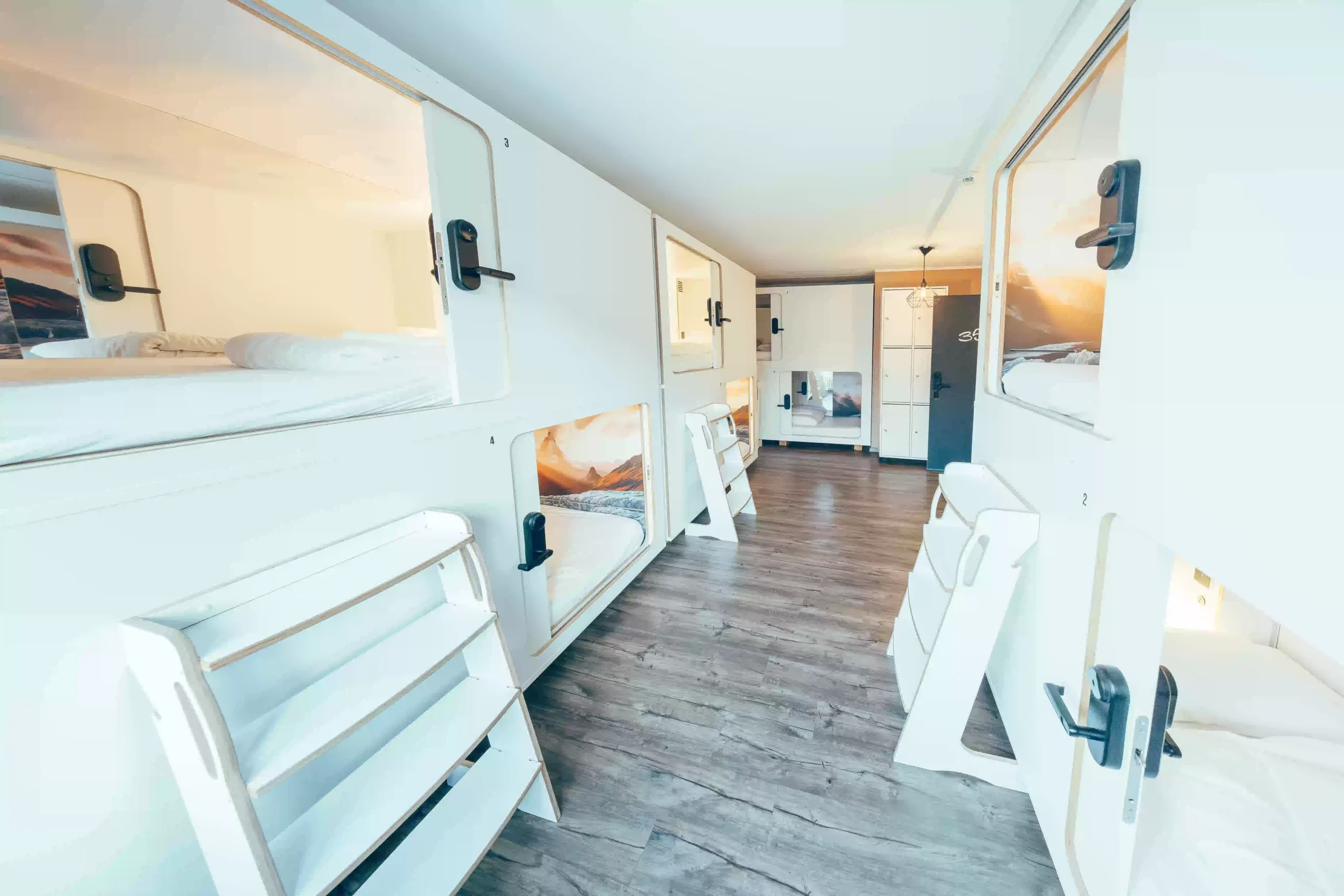 Bright dorm with double-row white sleeping pods, ladders, wood floors and lockers at Capsule Hotel