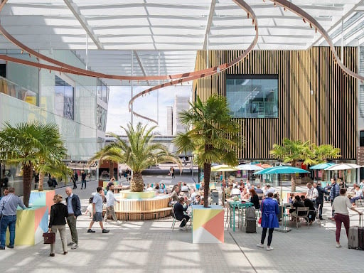 Capsule Hotel outdoor plaza with glass canopy, palm trees, cafe seating and guests socializing in a modern urban setting.